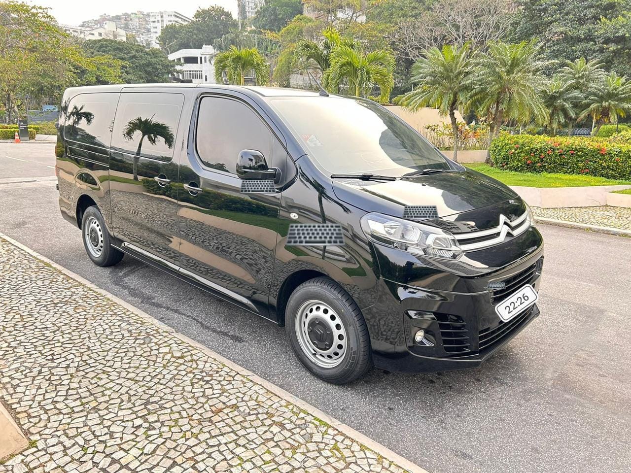 Van Citroën preta brilhante estacionada numa via com calçada de pedra portuguesa, palmeiras e prédios ao fundo, placa com tarja digital 22·26